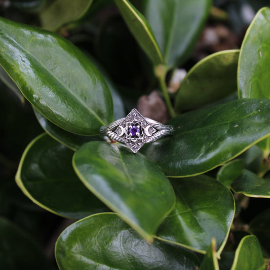 An engagement ring with a purple amethyst centerpiece, lotus flower design, and crescent moon detail, placed on a green leafy branch.