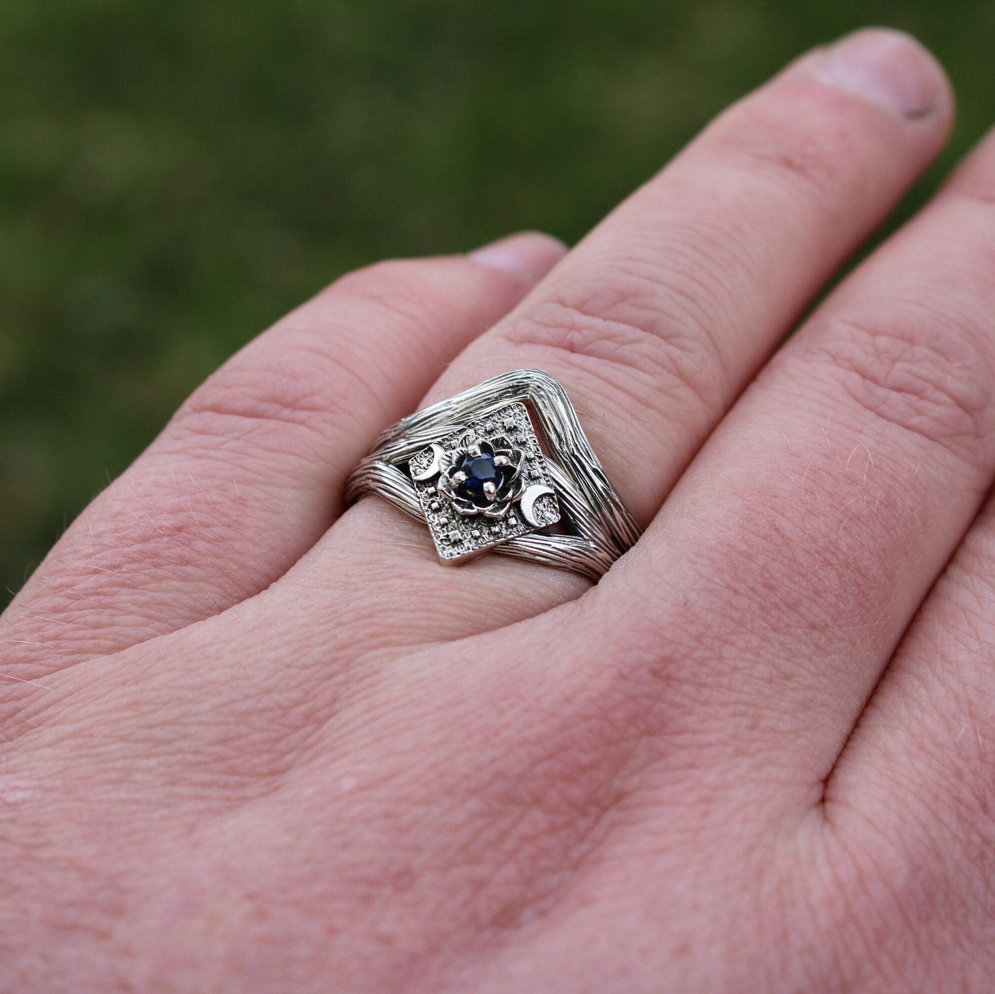 A close-up image of a person's hand displaying a sterling silver engagement ring with a sapphire center stone and lotus flower design, and a wedding band beside it.