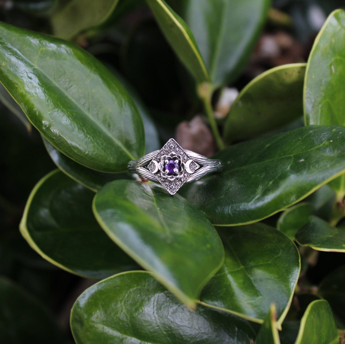 An engagement ring with a purple amethyst centerpiece, lotus flower design, and crescent moon detail, placed on a green leafy branch.