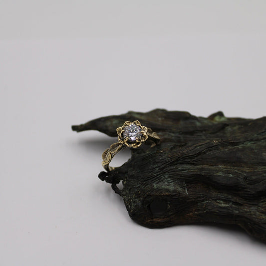 A 14k gold moissanite lotus engagement ring with a textured band and a central moissanite stone, displayed on a simulated natural branch.