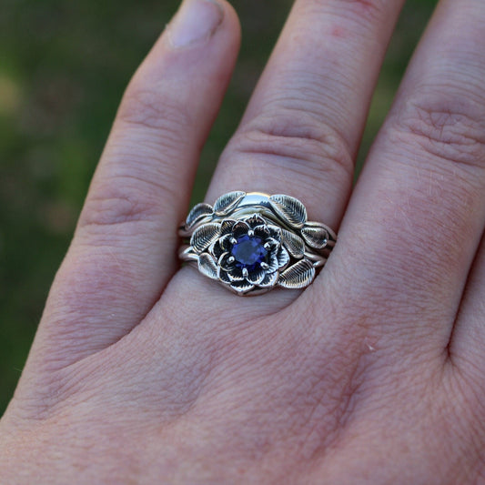 A close-up image of a sterling silver ring with an iolite stone set in a lotus design, held on a human hand.