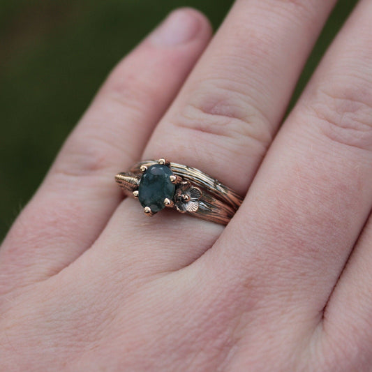 A close-up image of a hand displaying a gold ring with a large green moss agate stone and smaller clear stones set around it.