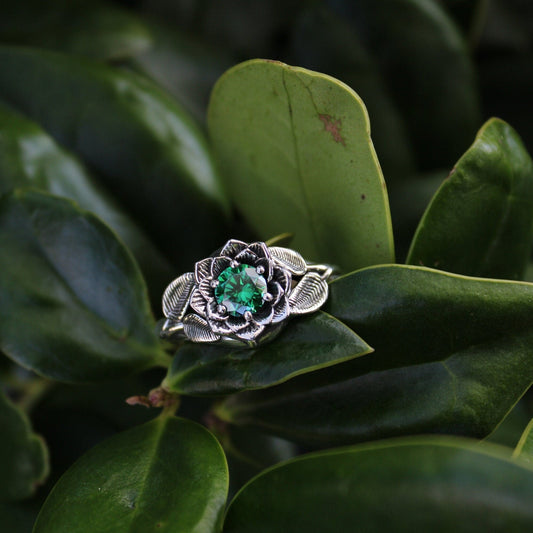 A sterling silver ring with a green cubic zirconia gemstone in a lotus design, placed on a leafy background.
