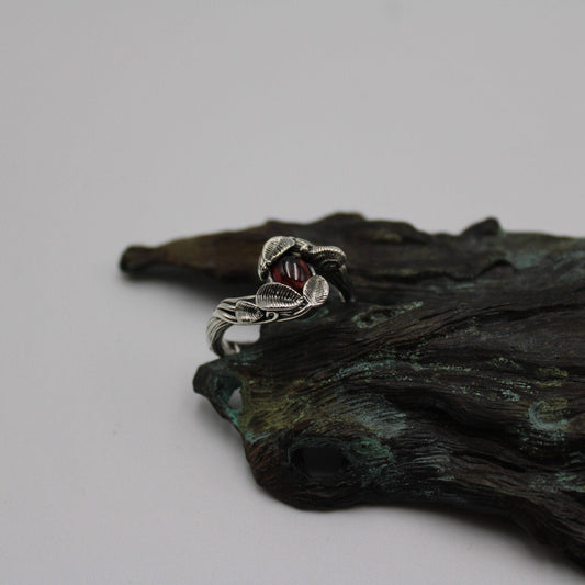 A sterling silver ring featuring a garnet gemstone, with intricate snail and leaf details, set on a textured silver band, displayed on a wooden surface.