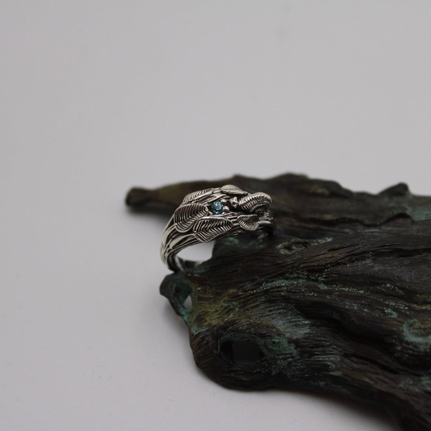 A sterling silver ring featuring a snail design with a blue topaz stone, displayed on a textured background that resembles a tree branch.