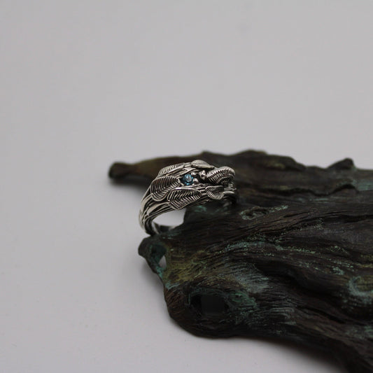 A sterling silver ring featuring a snail design with a blue topaz stone, displayed on a textured background that resembles a tree branch.